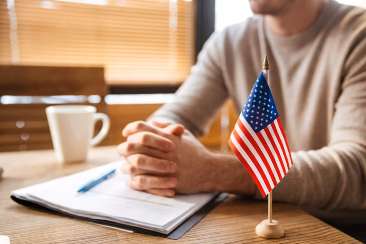 Primer plano de una persona llenando formularios de solicitud de visa de Estados Unidos, con un pasaporte abierto y una bandera de EE. UU. sobre el escritorio.